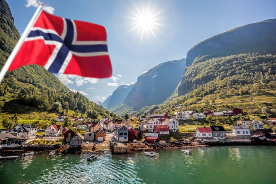 Natural Pharmaceuticals Movisan Norra küla fjordi ääres, mida ümbritsevad rohelised künkad ja järsud mäed päikesepaistelisel päeval. Esiplaanil lehvib tuules Norra lipp, taustaks rahulik siniroheline vesi ja maalilised majad.