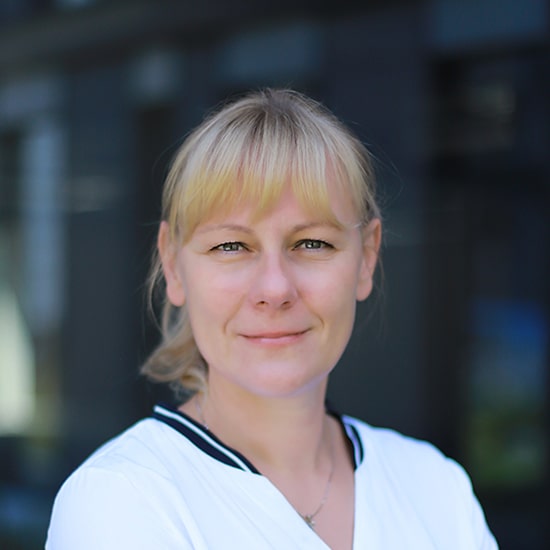 Natural Pharmaceuticals Movisan Blondide juuste ja hõredate karvadega naine, seljas valge särk, mille kaelus on tumedate kaunistustega, vaatab kaamerasse ja naeratab kergelt. Taustal on ähmased, moodsad, tumedad hooned.