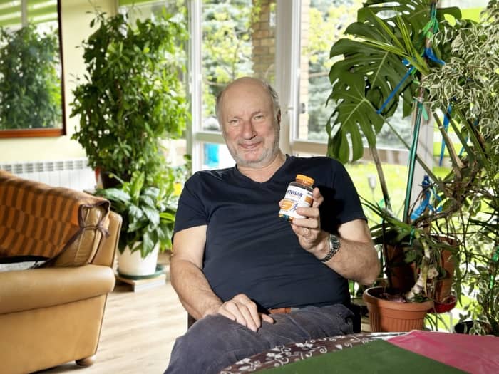 Natural Pharmaceuticals Movisan Eakas mees istub heledas, taimedega täidetud elutoas ja näitab naeratades Movisani toidulisandi pakendit. Taustal on näha rohelisi potitaimi, akent ja diivanit.