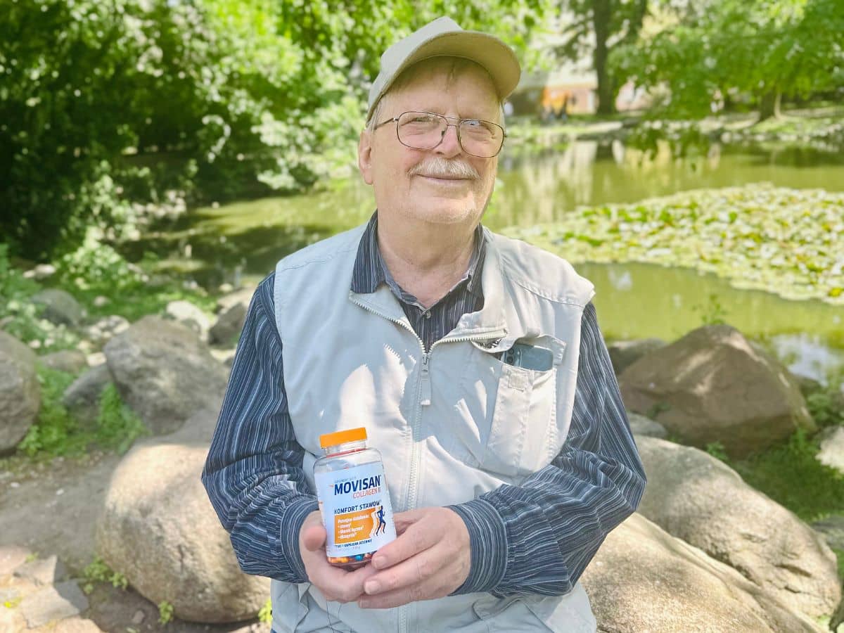 Natural Pharmaceuticals Movisan Prillidega, pesapallimütsi ja vestiga vanem mees istub kivil pargis tiigi ääres. Ta hoiab käes Movisan® Collageni toidulisandi pakendit, mida ta kaamerale näitab. Taustal on näha vesi koos vesirooside ja tiheda rohelusega.
