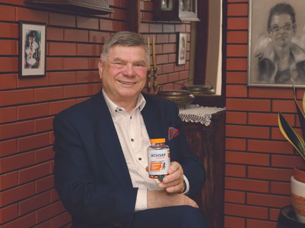 Natural Pharmaceuticals Movisan Vanem ülikonnas mees istub elutoas vastu punast telliskiviseina ja hoiab käes pakendit toidulisandiga MOVISAN.